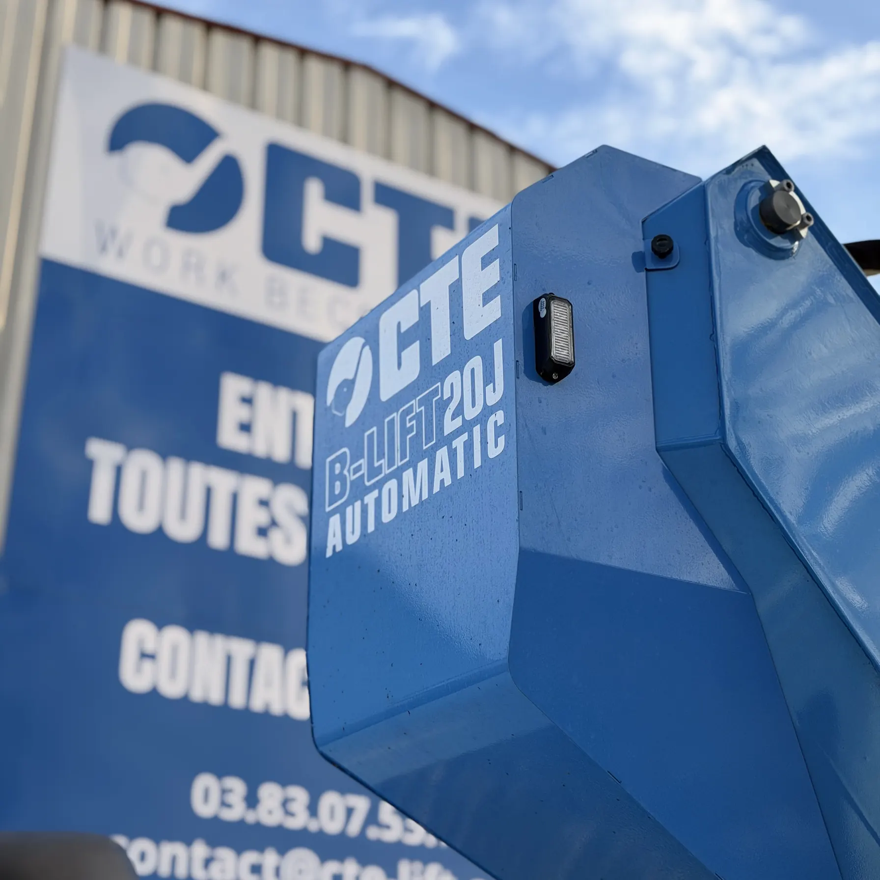 Gros plan sur la flèche bleue de la nacelle marquée CTE B-LIFT AUTOMATIC, devant l'enseigne de l'atelier CTE Lift